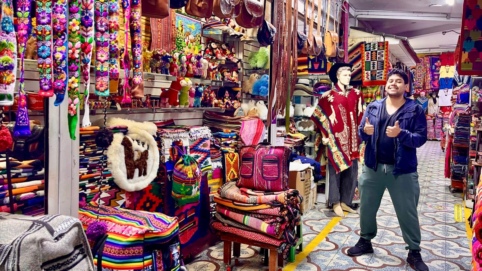 Diego Rios stands in a vibrant traditional souvenir market in Pueblo Libre, Lima, surrounded by colorful Peruvian textiles and handicrafts. Kieran Proctor is behind the camera, taking the photo.